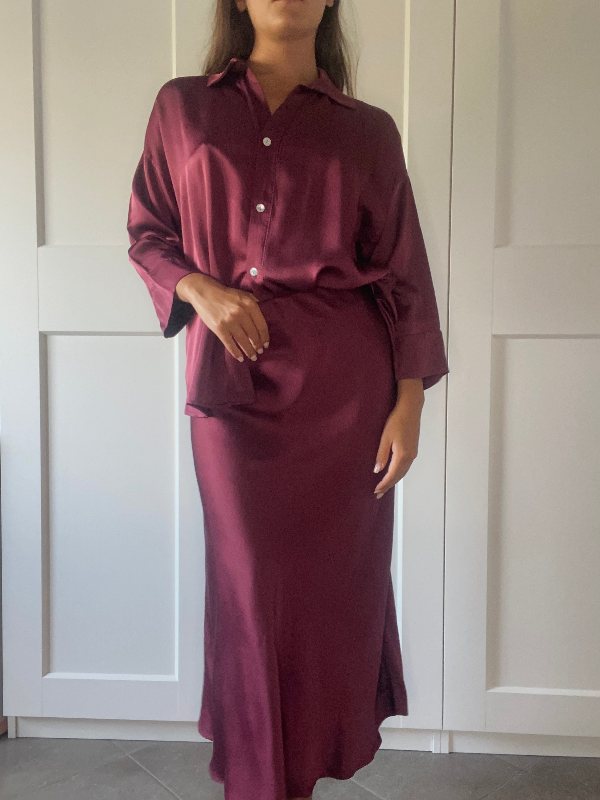 Donna che indossa un completo in seta bordeaux, composto da camicia e gonna lunga, posando davanti a porte bianche.
