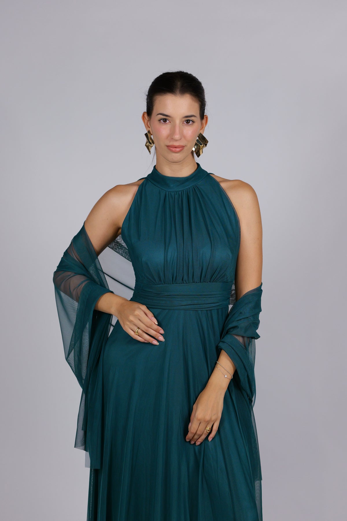 Donna con abito elegante di colore verde scuro, collo alto a imbarcatura, gonna lunga, con foulard in tulle trasparente, su sfondo chiaro.
