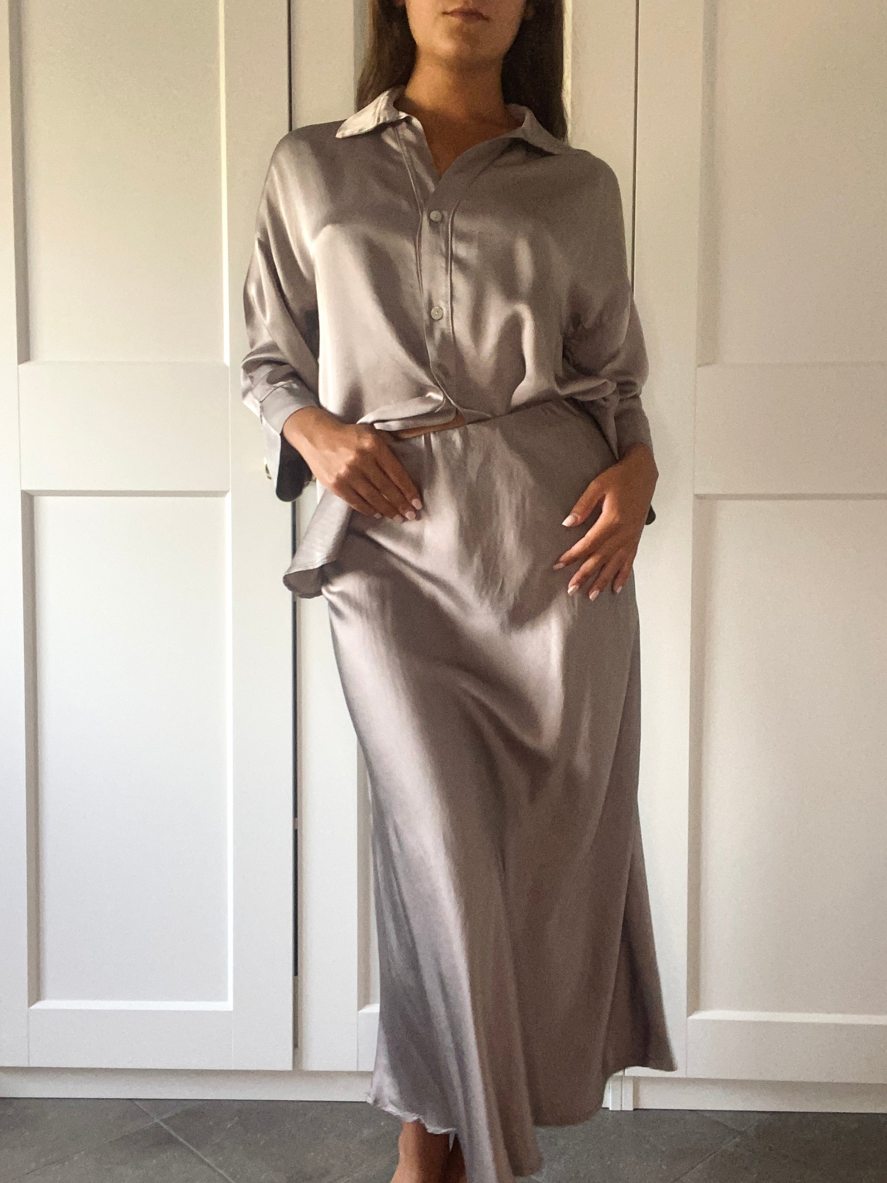 Donna con capelli lunghi indossa un completo in seta beige, camicia e gonna, sfondo con porte bianche.
