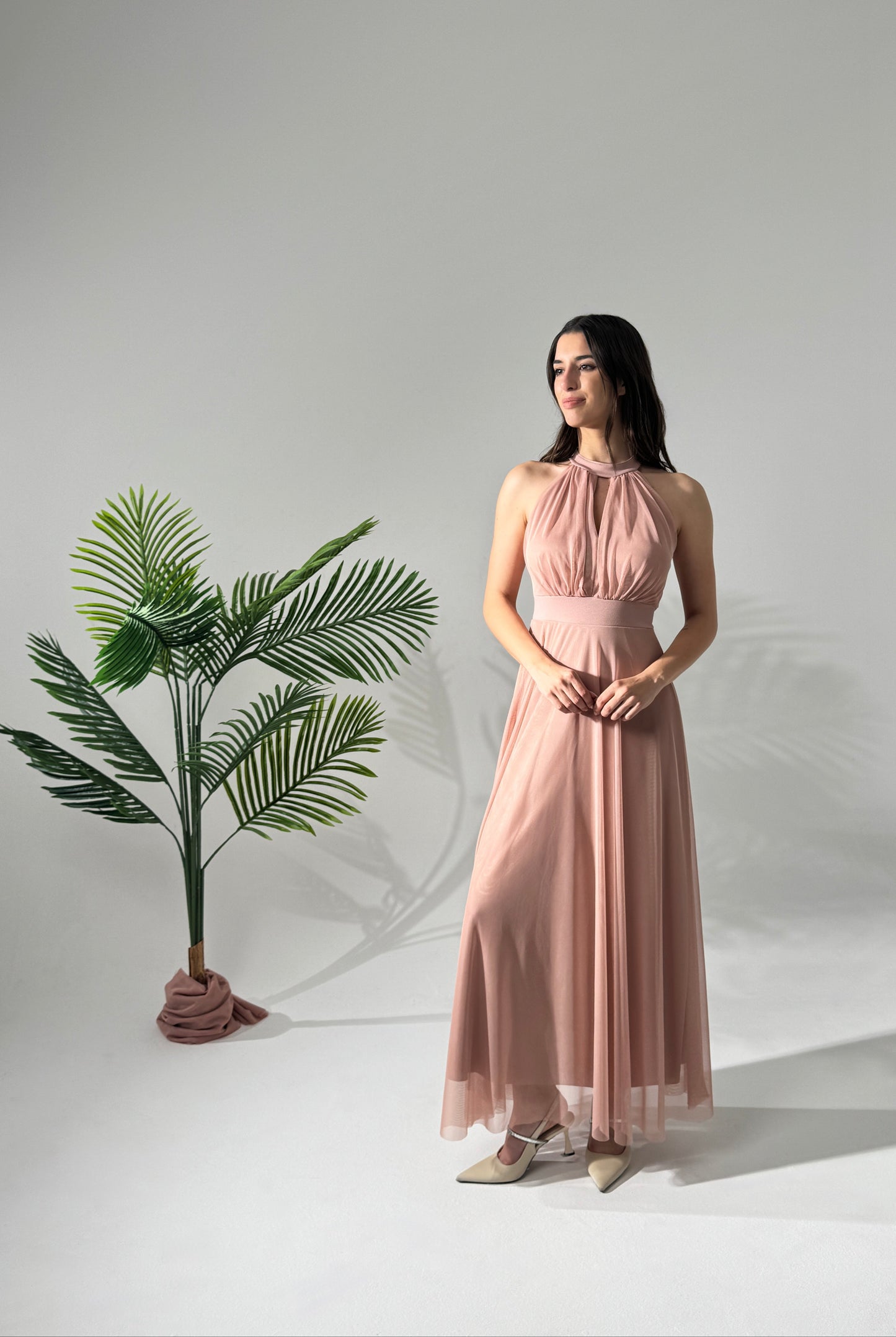 Donna con abito lungo rosa, collo alto a nodo, accanto a pianta verde