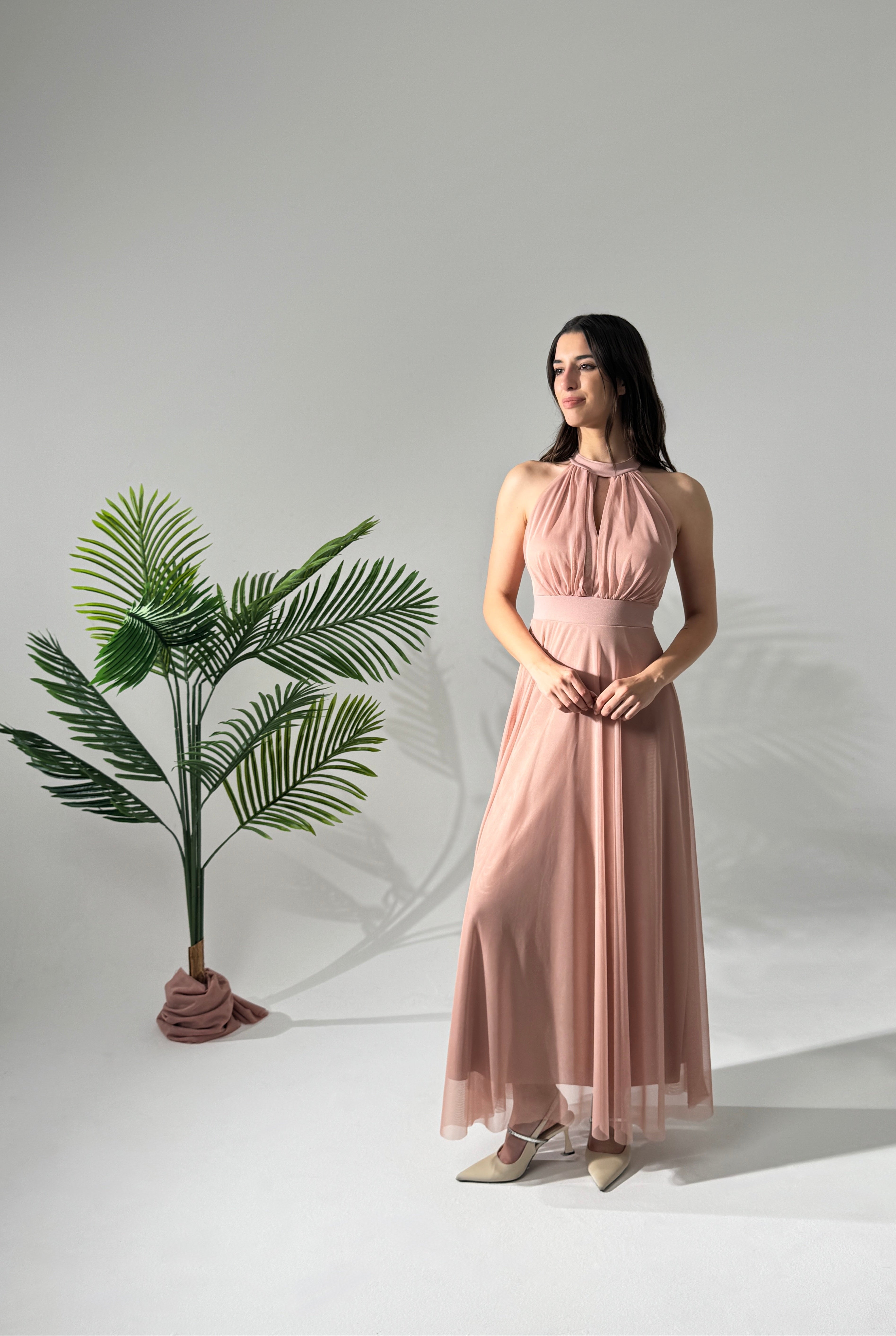 Donna con abito lungo rosa, collo alto a nodo, accanto a pianta verde
