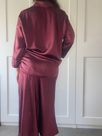  Outfit da donna in seta bordeaux: camicia oversize a maniche lunghe e gonna maxi, posata davanti a porte bianche.
