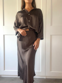 Outfit elegante in seta fango: camicia over e gonna maxi, indossato da una donna davanti a porte bianche.
