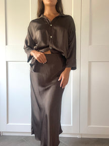 Outfit elegante in seta fango: camicia over e gonna maxi, indossato da una donna davanti a porte bianche.
