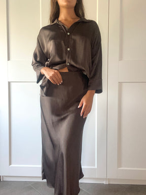 Outfit elegante in seta fango: camicia over e gonna maxi, indossato da una donna davanti a porte bianche.
