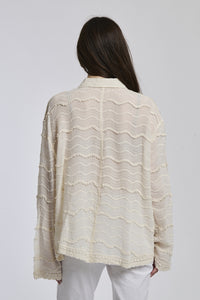 Camicia a maniche lunghe beige con texture ricamata, abbinata a pantaloni bianchi, sfondo bianco