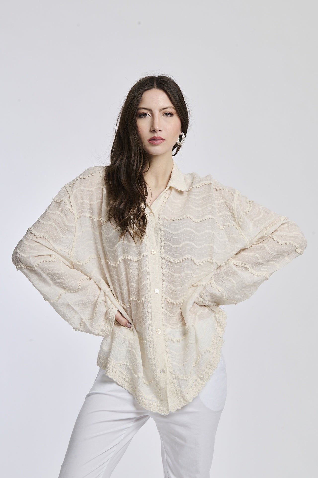Donna con camicia oversize in pizzo beige e pantaloni bianchi, posa con mani sulle fianche