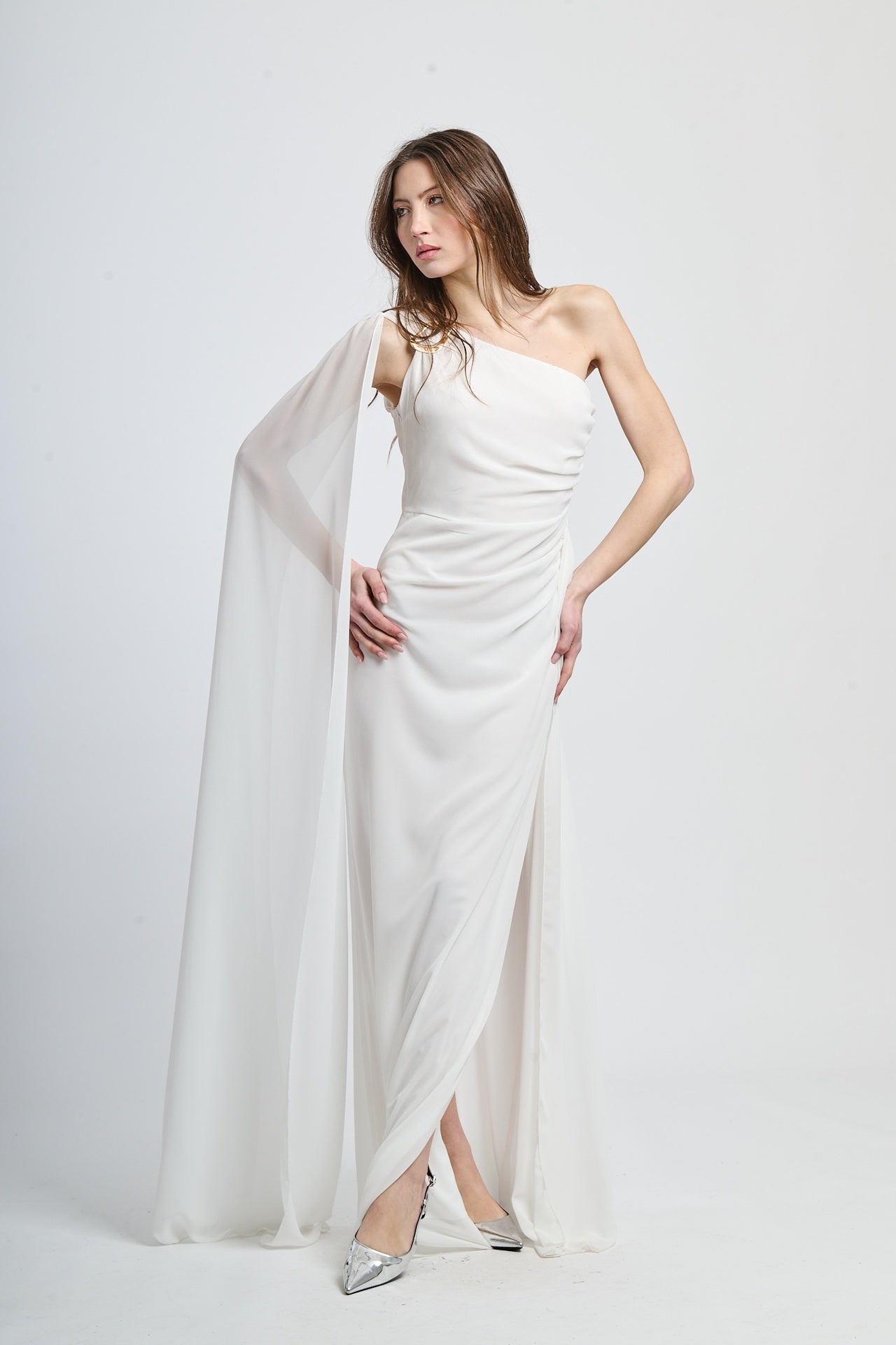 Abito lungo bianco, spalla singola, drappeggio fluido, accessorio decorativo sulla spalla.