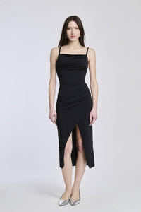 Donna con abito midi nero, taglio aderente, spalline sottili e sfilo che crea un’effetto drammatico.