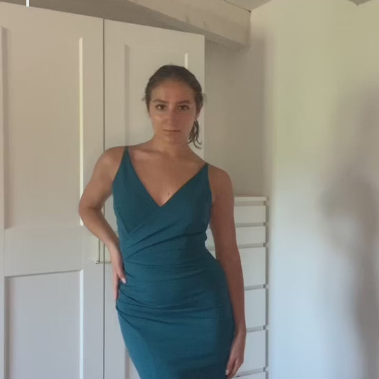  donna indossa un abito elegante blu scuro con spalline sottili e uno sguardo accattivante, posa in uno spoglio interno con armadio bianco sullo sfondo