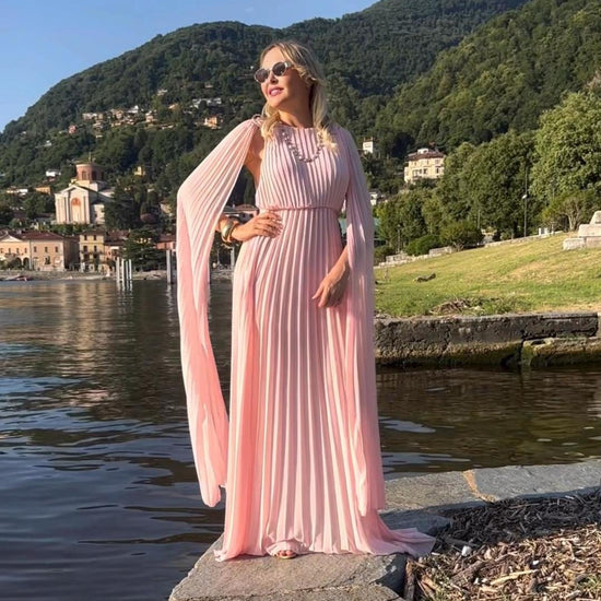 Donna con abito rosa plissé e mantelle, posa sul lago con montagne sullo sfondo